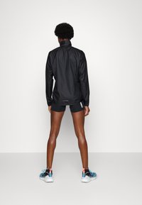 Persona de espaldas, vistiendo una chaqueta ligera negra, pantalones cortos negros y zapatillas deportivas azules y rosas sobre un fondo simple.