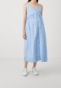 Ljusblå midi-klänning med justerbara band, knappar fram, stjärnmönster broderi och detaljerad kantdekoration. Kombineras med vita sneakers.