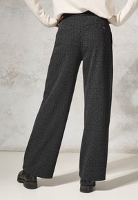 Široké kalhoty z texturované černé tkaniny s jemným vzorem. Mají vysoký pas a malou zadní kapsu, stylizované s kotníkovými botami.