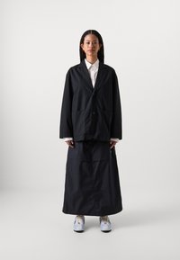 Svart, oversized jakke med to frontlommer, lagd over en matchende lang skjørt. Hvit skjorte med krage under og lyseblå sneakers.