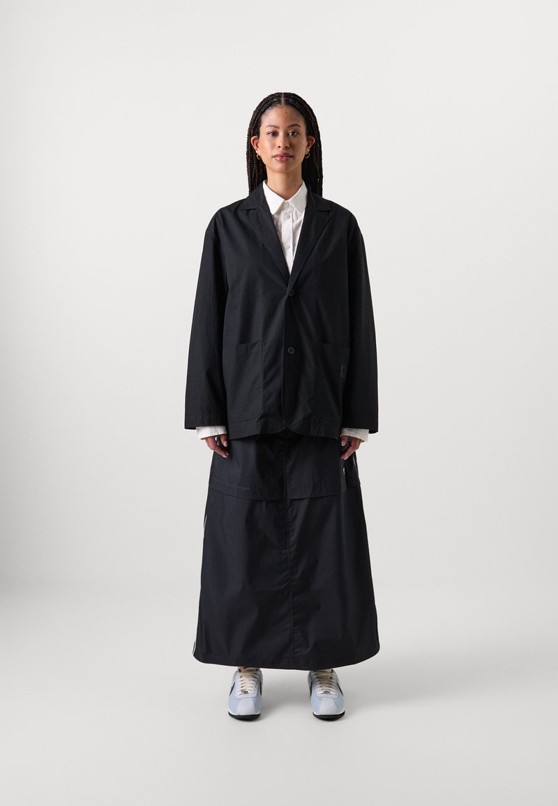 Svart, oversized jakke med to frontlommer, lagd over en matchende lang skjørt. Hvit skjorte med krage under og lyseblå sneakers.