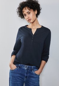 Marineblaues, langärmliges Oberteil mit Henley-Ausschnitt und drei Knöpfen; weicher Stoff; kombiniert mit blauen Jeans im klassischen Schnitt.