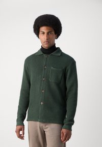 Cardigan de punto texturizado verde oscuro con cuello, que presenta cinco botones de madera y un bolsillo en el pecho, combinado con pantalones beige.