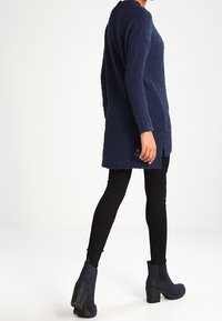 Une femme porte un long pull texturé bleu marine, des leggings noirs et des bottines bleu marine à talons épais, debout devant un fond blanc.