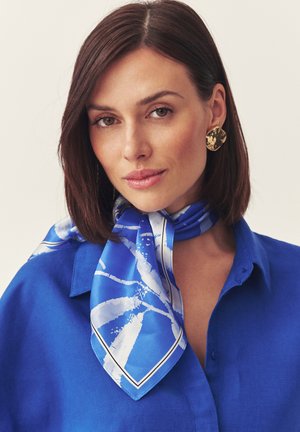Femme aux cheveux bruns portant une chemise bleue, un foulard en soie bleu et blanc à motifs noué autour du cou, et de grandes boucles d’oreilles dorées texturées.