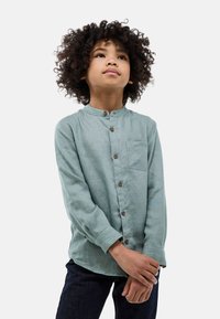 Chemise vert clair à col mandarin, à manches longues et avec une poche avant. Dotée de boutons marron et d'un tissu doux et texturé.