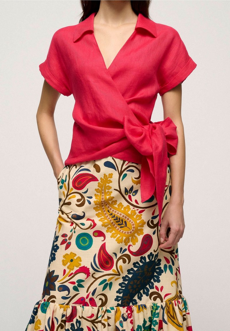 Top avvolgente rosso in lino con maniche corte e un dettaglio a fiocco in vita, abbinato a una gonna color crema con motivi paisley e floreali colorati.