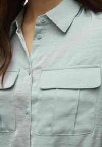 Camicia button-up azzurro chiaro con colletto, due tasche sul petto e texture liscia. Presenta un tessuto morbido e bottoni piccoli per la chiusura.