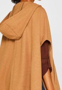 Mujer que lleva una capa con capucha color beige sobre una camiseta de manga larga marrón, mostrando un brazo por una abertura de la capa.