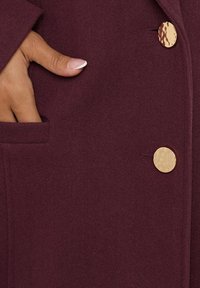 Burgundy takki, jossa on tekstuuripinta, sivutasku ja yksittäinen kultasävyinen hakasnapin kiinnitys.