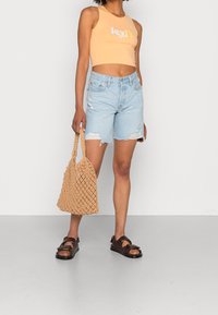 Lososová zkrácená tílko s bílým logem, světlé modré oprané džíny, tkaná béžová taška a hnědé sandály s dvojitými p ásky.