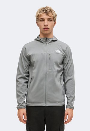 Junger Mann mit kurzen blonden Haaren, trägt eine graue Kapuzenjacke mit Reißverschluss und schwarze Hose, steht vor einem schlichten Hintergrund.