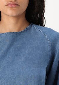 Svjetloplavi traper majica s okruglim izrezom, raglan rukavima i mekom teksturom. Ima suptilni sjaj i šivane detalje.