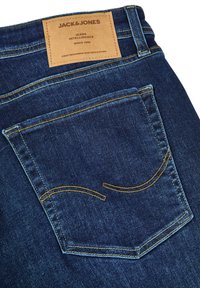 Jeans de mezclilla en azul oscuro con costuras naranjas. Cuenta con un bolsillo trasero y una etiqueta beige con el nombre de la marca y detalles cosidos en la cintura.