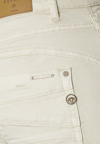 Los vaqueros de mezclilla de color crema claro presentan un primer plano de paneles cosidos, detalles metálicos y una etiqueta de cuero con la marca en la cinturilla.