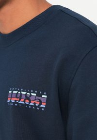 Pogled z bližine na osebo, oblečeno v temno modro majico s kapuco z napisom "Ustanovljeno 1985 Tommy Jeans" in barvno črtasto grafiko na prsih.