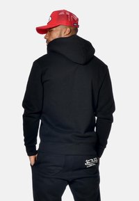 Uomo che indossa un hoodie nero e pantaloni con logo bianco sulla tasca posteriore, cappellino da baseball in rete rossa, rivolto di spalle su sfondo semplice.