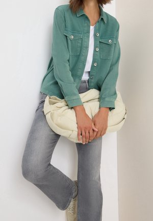 Person iført en grøn knappet jakke, hvid skjorte og grå jeans, holder en stor cremefarvet blød taske, mens de læner sig op ad en væg.