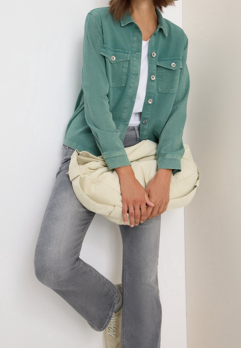 Personne portant une veste verte boutonnée, une chemise blanche et un jean gris, tenant un grand sac doux couleur crème tout en s'appuyant contre un mur.