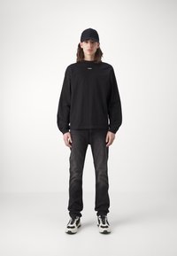 Black long-sleeve shirt with a small logo, paired with dark jeans and black-and-white sneakers. A black cap completes the outfit.