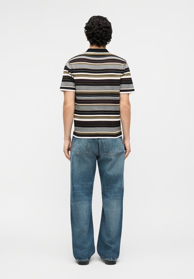 Άνδρας με κατσαρά μαλλιά, που φοράει κοντομάνικο πολύχρωμο ριγέ πουκάμισο και άνετο μπλε τζιν, στέκεται γυρνώντας την πλάτη σε ένα απλό φόντο.