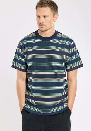 Homme portant un t-shirt bleu marine à manches courtes avec des rayures horizontales vertes, beiges et bleu clair, debout, mains dans les poches, regardant à gauche.