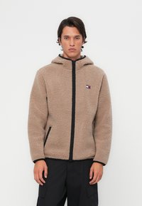 Giacca in pile marrone con cappuccio, dotata di chiusura zip nera e tasche laterali. Piccola patch logo sul petto sinistro. Tessuto morbido.