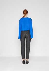 Blusa azul de manga larga con detalles plisados en la parte posterior, combinada con pantalones negros ajustados y zapatos negros, vista desde atrás.
