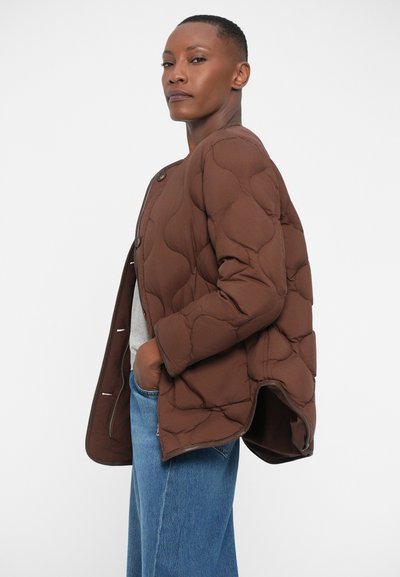 Giacca trapuntata marrone con una texture gonfia, grandi bottoni e tasche laterali, abbinata a jeans blu a gamba larga e una maglietta grigia chiara.