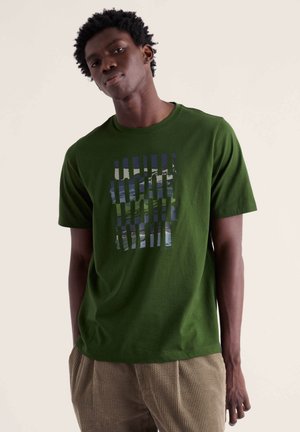Jeune homme portant un t-shirt vert avec un motif abstrait en grille bleu et blanc, associé à un pantalon beige en velours côtelé, debout devant un fond neutre.