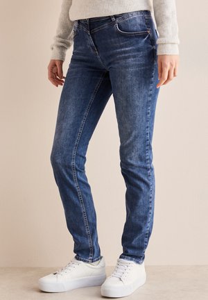 Dark blue denim jeans with a slim fit, five-pocket design, visible stitching, and worn-in texture, paired with white low-top sneakers.