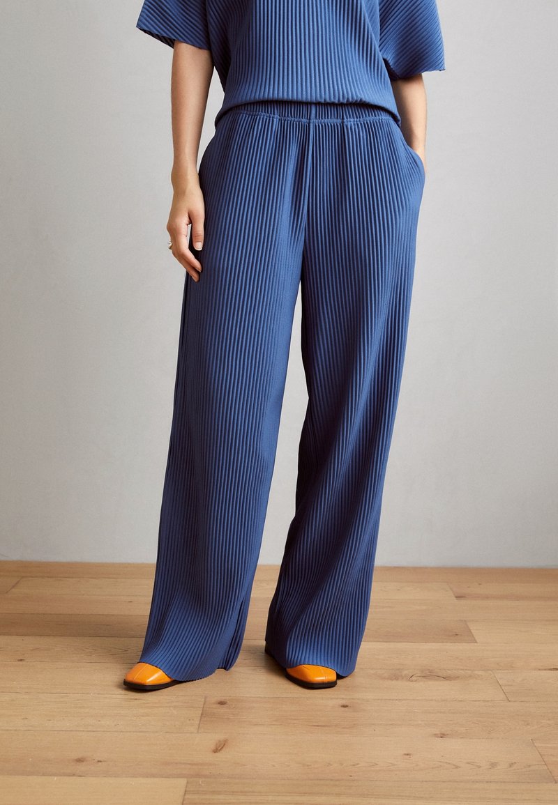 Femme en pantalon large bleu côtelé ample et top assorti, debout avec une main dans la poche, portant des chaussures orange vif sur un sol en bois.