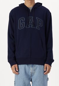 Sudadera con cremallera en color navy, hecha de una mezcla de algodón, que incluye bolsillos frontales, un gran logo "GAP" en azul oscuro y capucha con cordón. Textura suave.