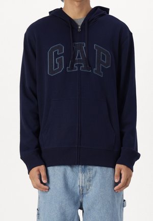 Sudadera con cremallera en color navy, hecha de una mezcla de algodón, que incluye bolsillos frontales, un gran logo "GAP" en azul oscuro y capucha con cordón. Textura suave.