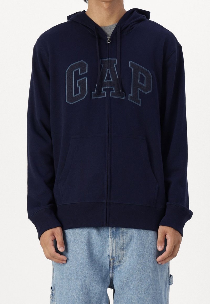 Sudadera con cremallera en color navy, hecha de una mezcla de algodón, que incluye bolsillos frontales, un gran logo "GAP" en azul oscuro y capucha con cordón. Textura suave.