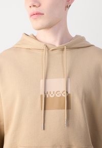 Felpa beige con cordini, caratterizzata da un logo rettangolare in marrone chiaro e scuro con il testo "HUGO". Tessuto di cotone dal tocco casual.
