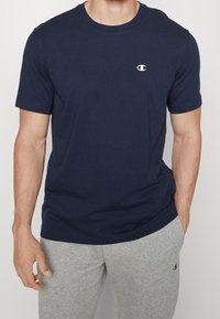 Maglietta in cotone blu navy con scollatura rotonda e maniche corte. Presenta un piccolo logo bianco sul petto. Abbinata a pantaloni della tuta grigi.