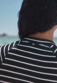 Persona con capelli scuri che indossa un maglione a righe bianche e nere con trama, vista da dietro con uno sfondo marino sfocato.