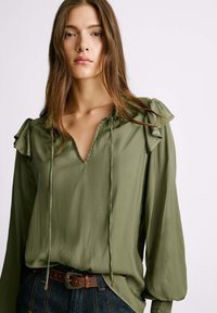 Blusa verde oliva con maniche lunghe, dettagli a balze, scollatura a V e decorazioni in tono oro lungo il colletto. Tessuto dalla texture liscia.