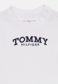 Tricou alb din bumbac, cu text brodat de culoare albastră marină care spune "TOMMY HILFIGER" pe piept, cu nasturi la umăr.