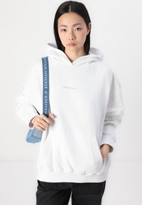Hvit hettegenser med frontlomme, med «PEGADOR» logo. Kombinert med mørke jeans og en blå crossover-veske. Glatt tekstur.