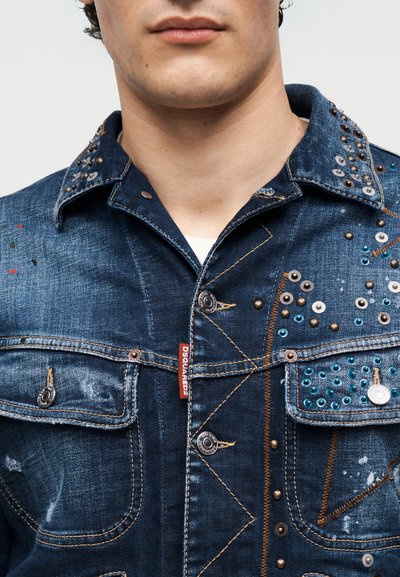 Άνδρας που φοράει σκούρο μπλε τζιν μπουφάν με μεταλλικά καρφιά, οπές, πορτοκαλί ραφές και κόκκινη ετικέτα Dsquared2 στην μπροστινή τσέπη.
