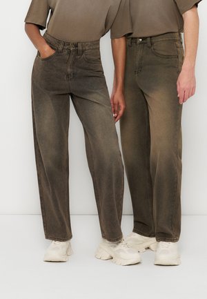 Kaks inimest, kes kannavad pleekinud pruune sirge säärega pükse, beeže toppe ja valgeid paksu tallaga tossusid, seisavad lameda valge tausta ees.