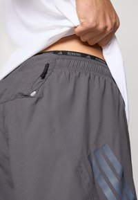 Harmaat juoksushortsit, joissa on joustava vyötärönauha, jossa teksti "RUNNING", sivutaskut ja heijastava kolmiviivainen kuvio jalassa.