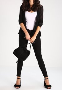 Cardigan negro de punto superpuesto sobre una camiseta blanca, combinado con jeans skinny negros y sandalias de tacón negro con acentos de volantes. Lleva un bolso negro.