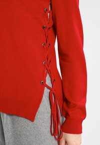 Pull en tricot rouge avec un détail de laçage sur les côtés et des œillets en métal. Présente une coupe décontractée et un tissu texturé. Associé à un bas gris.