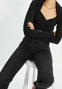Top nero a maniche lunghe con scollatura quadrata e arricciature; abbinato a jeans in denim scuro a vita alta; seduta su uno sgabello bianco.