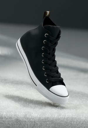Czarny sneaker typu high-top z białym czubkiem i podeszwą, czarnymi sznurowadłami oraz pasiastą pętlą z tyłu, unoszący się nad miękką, teksturowaną powierzchnią.