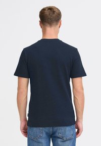 Tamsiai mėlyna trumpomis rankovėmis marškinėliai su tekstūruotu audiniu, apvalia iškirpte ir maža prekės ženklo etikete šone. Vaizdas iš nugaros.