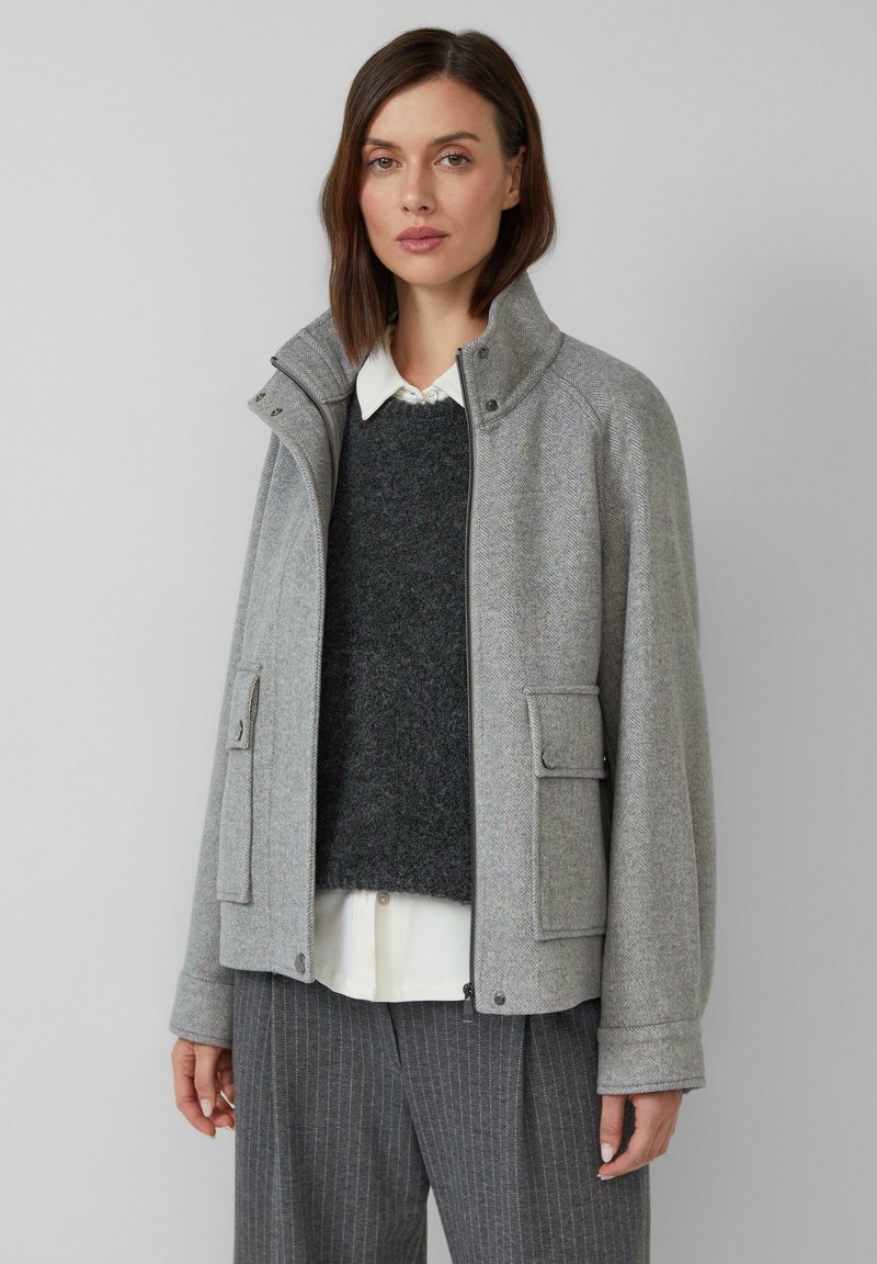 Grau melierte Jacke aus Wollmischung mit Fischgrätenmuster, Reißverschluss vorne, hohem Kragen und zwei Fronttaschen. Getragen über einem dunklen Pullover und einem weißen Hemd.